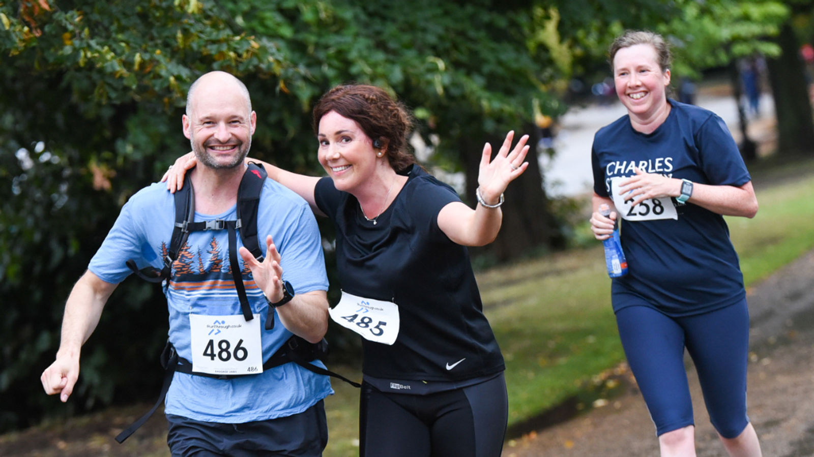 Three people jogging along a path during a race. The man on the left wears a numbered bib and a backpack, smiling. The woman in the middle also wears a bib, with both arms outstretched. Another woman follows behind, holding a water bottle, also smiling.