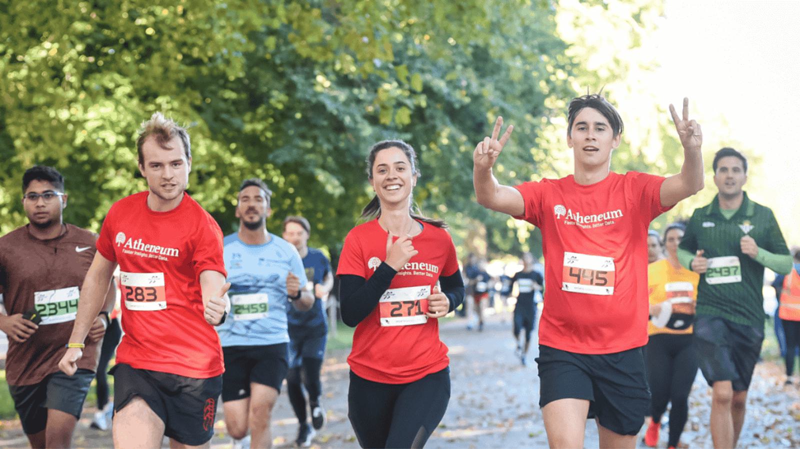 A group of runners participate in a race on a tree-lined path. Three people in red shirts lead, smiling and making gestures. Other runners in various colored outfits follow. The scene is vibrant and energetic under a clear sky.