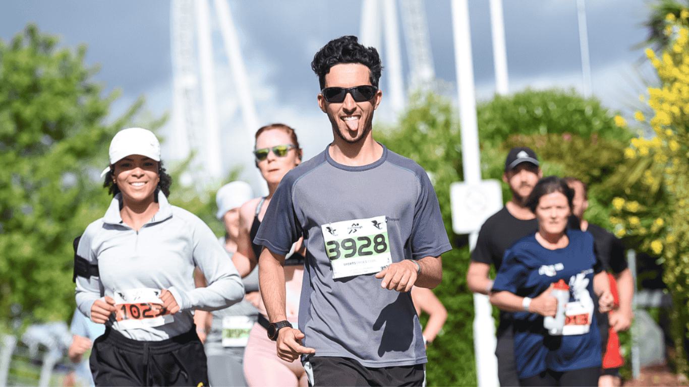 A group of runners participating in a race, with a man in a gray shirt and sunglasses leading the pack, smiling and sticking out his tongue. The background shows green trees and a sunny sky.