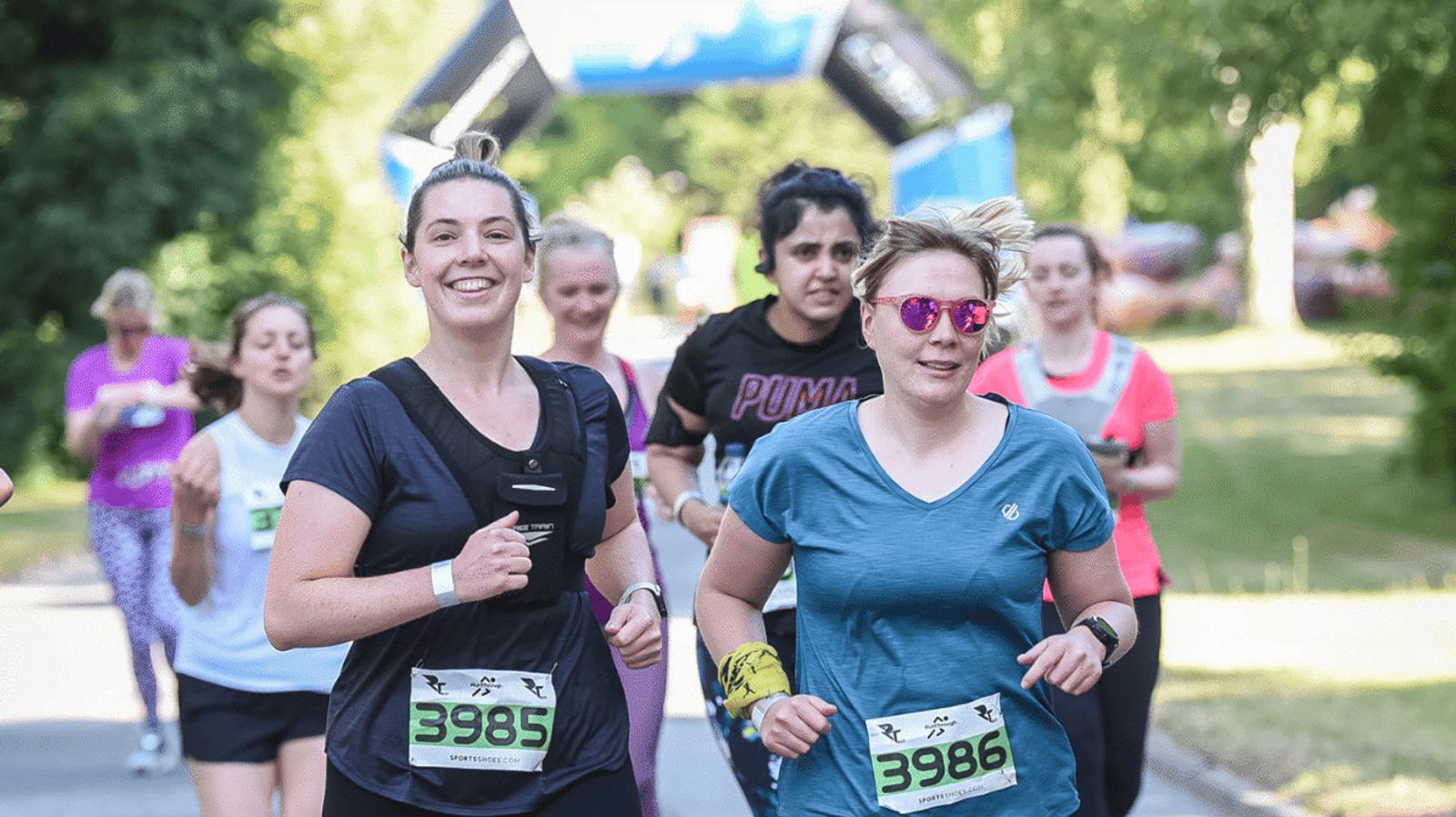 A group of runners participating in a race on a sunny day. Two women in the foreground are smiling and wearing numbered race bibs. Other runners are visible in the background. The path is lined with trees.