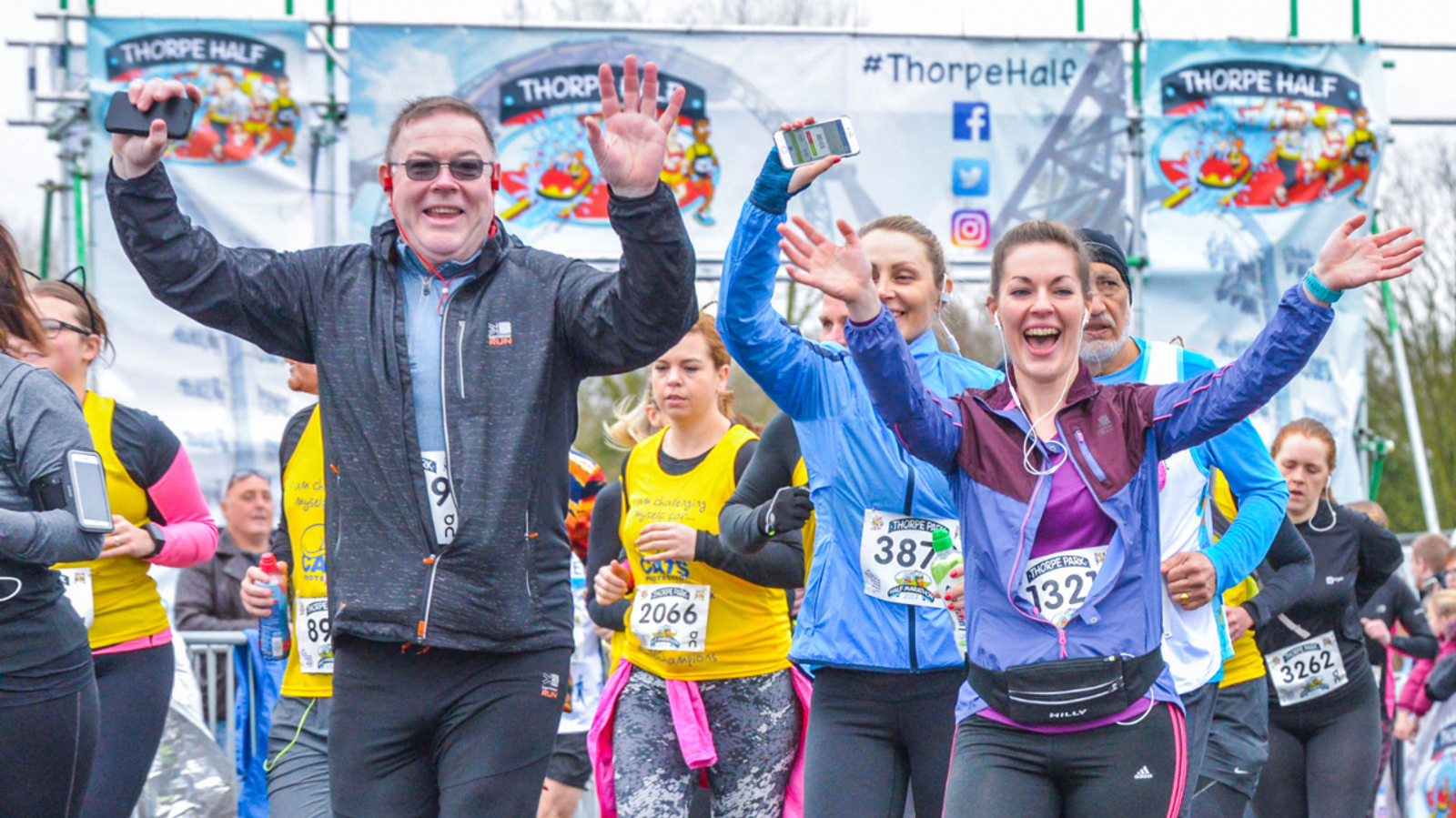 Runners happily waving as they cross the starting line of the Thorpe Half Marathon. Participants display their race numbers, and some hold phones. A banner with "Thorpe Half" is visible in the background.