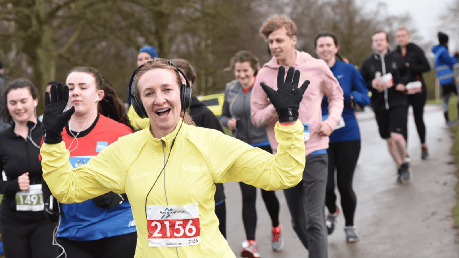 A group of runners participates in a race on a cloudy day. A smiling woman in a yellow jacket, wearing headphones, waves at the camera. She is surrounded by other participants, some wearing numbered race bibs. Trees line the background.
