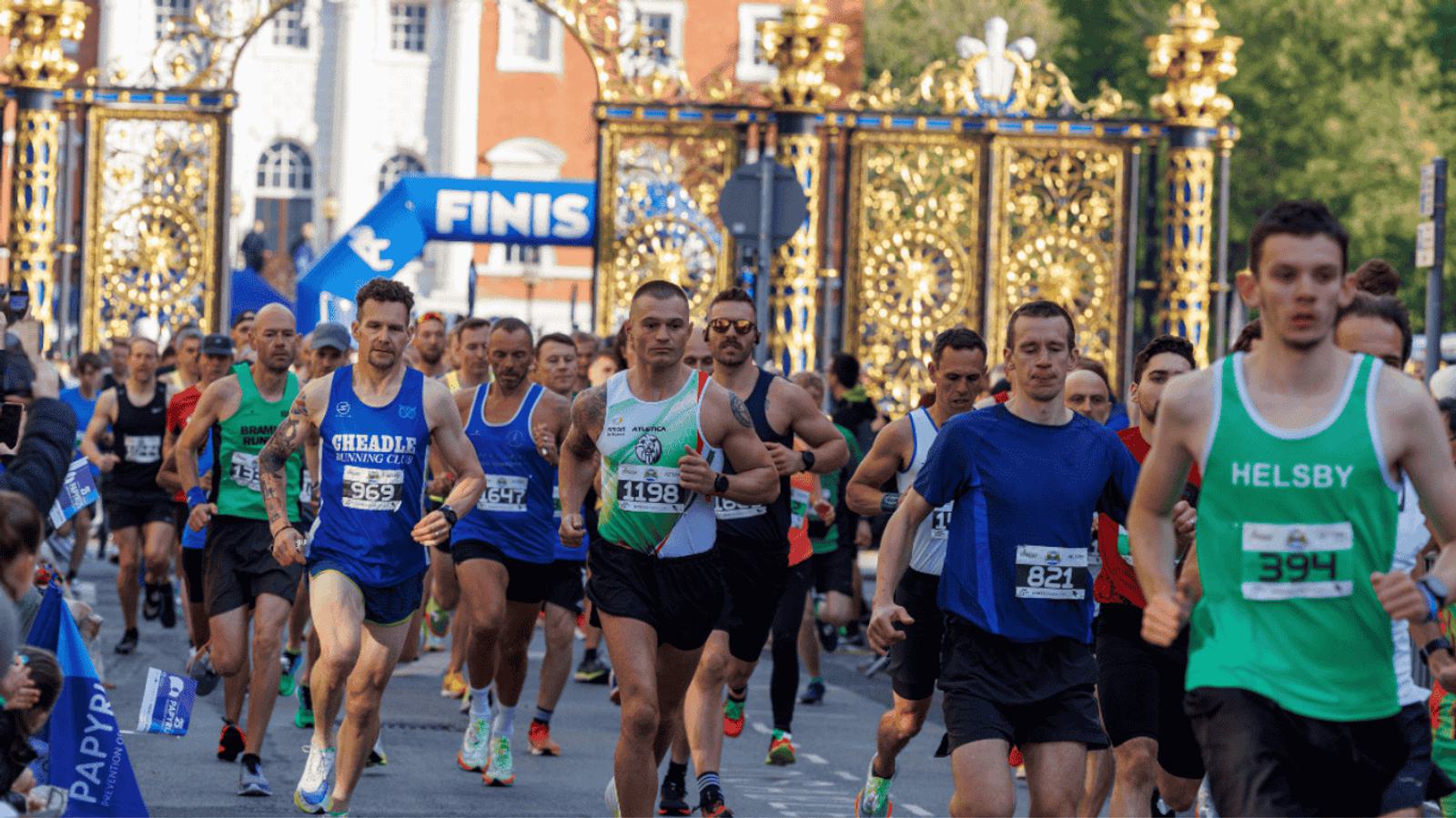 A group of runners nearing the finish line during a race. Participants wear various athletic gear, and a "FINISH" sign is visible in the background, with ornate gates and greenery behind them.