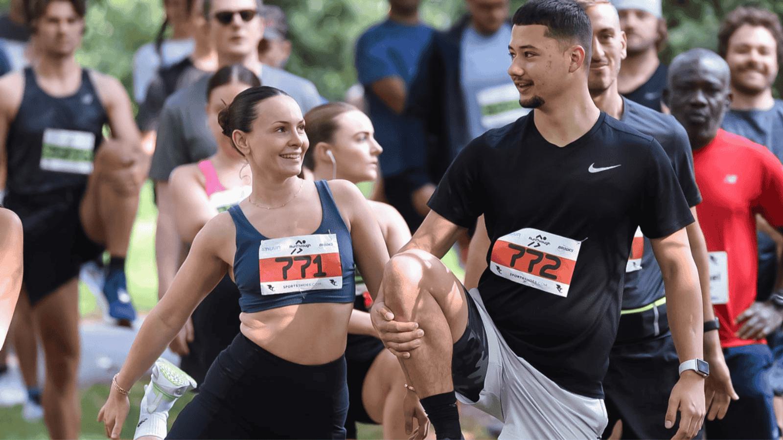 A group of people warming up before a run, with two individuals in the foreground stretching their legs and smiling at each other. They are outdoors, surrounded by trees, wearing running attire and bibs.