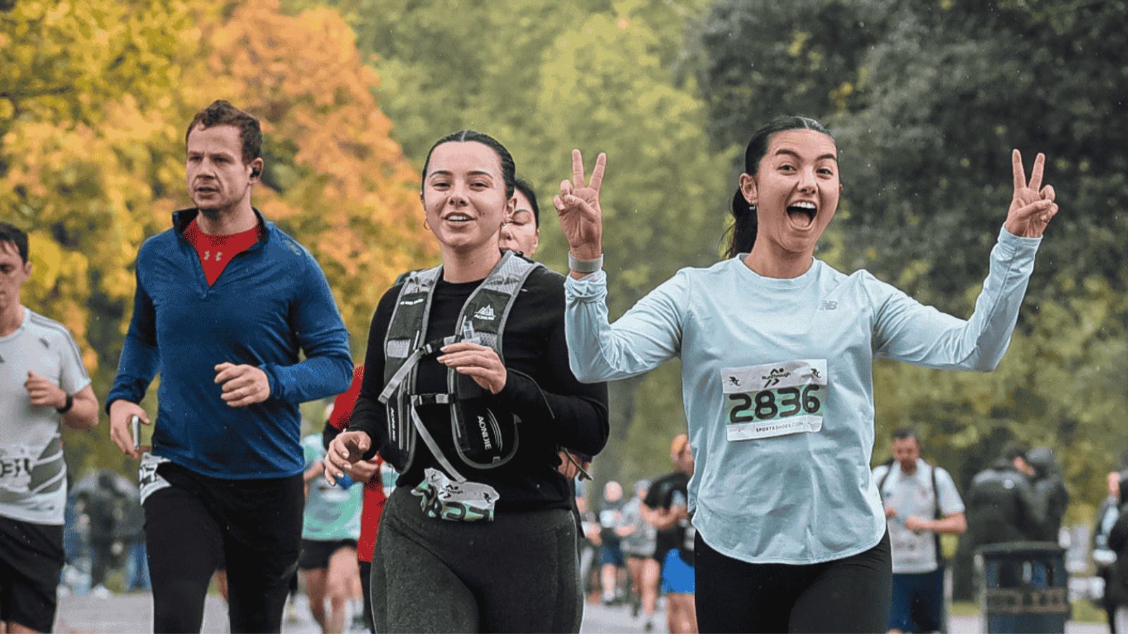 People running in a race through a park, with two women in the foreground smiling and making peace signs. They wear athletic gear and race bibs. Autumn trees are visible in the background, adding colorful foliage.