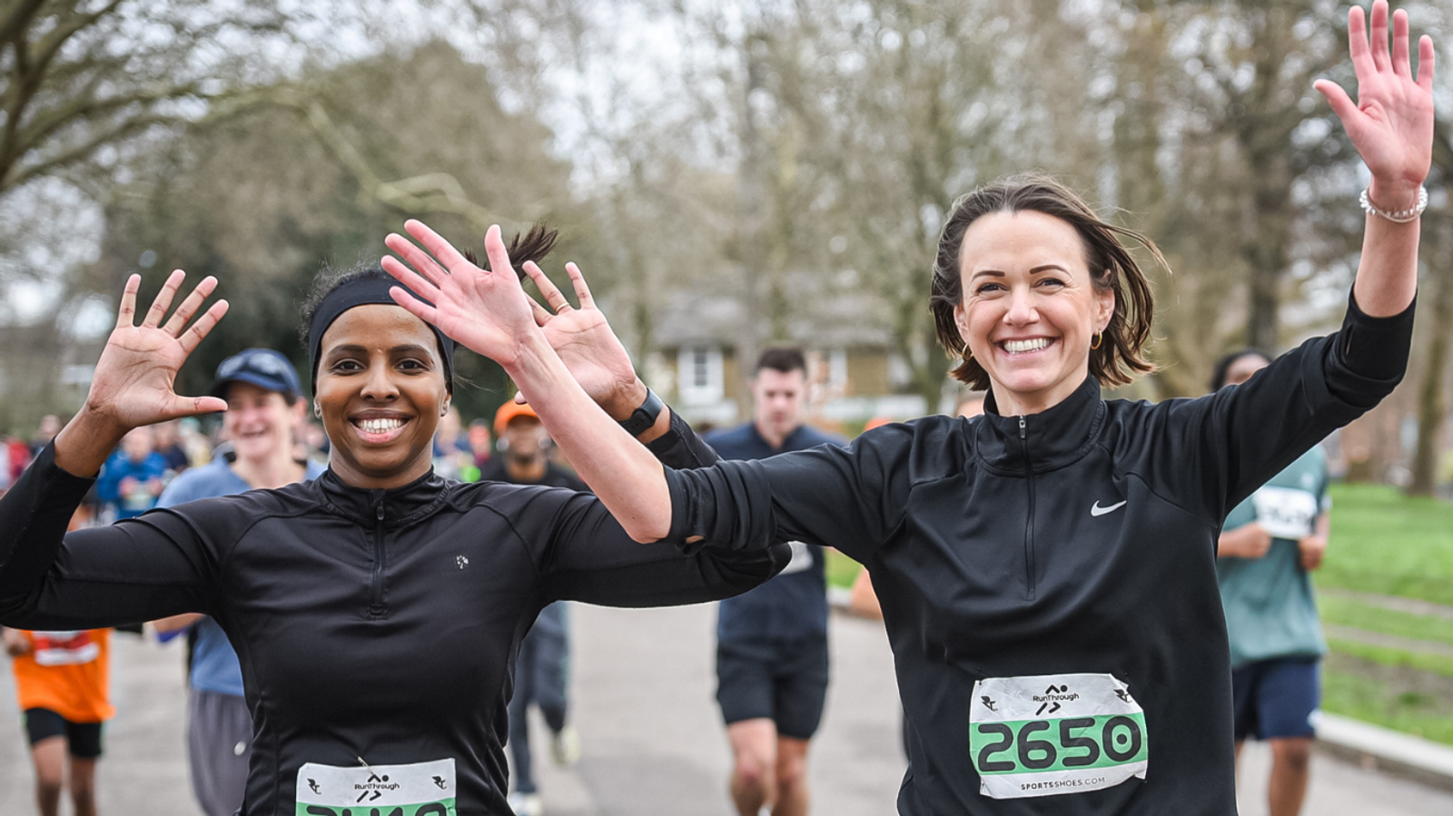 Two women smiling and running in a race, wearing black athletic outfits and race numbers. They are raising their arms in celebration. Other runners and trees are visible in the background.