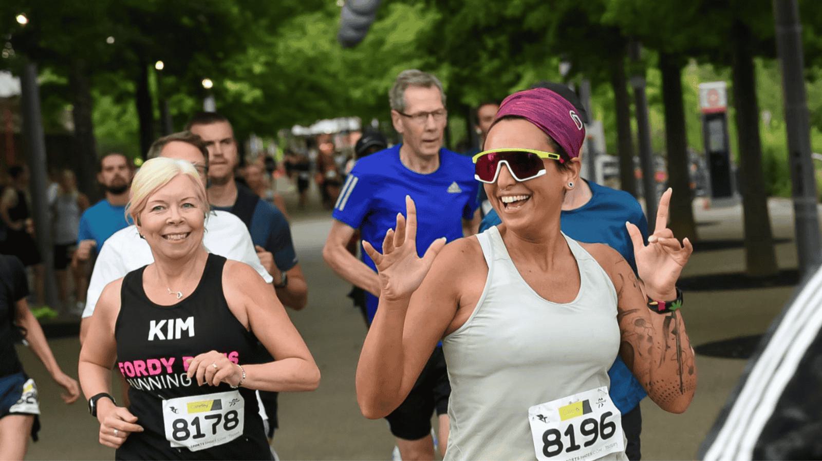 A group of runners participating in a race, with bib numbers visible. One woman in the foreground is smiling and gesturing with her hands. Lush green trees line the path, contributing to the lively outdoor atmosphere.