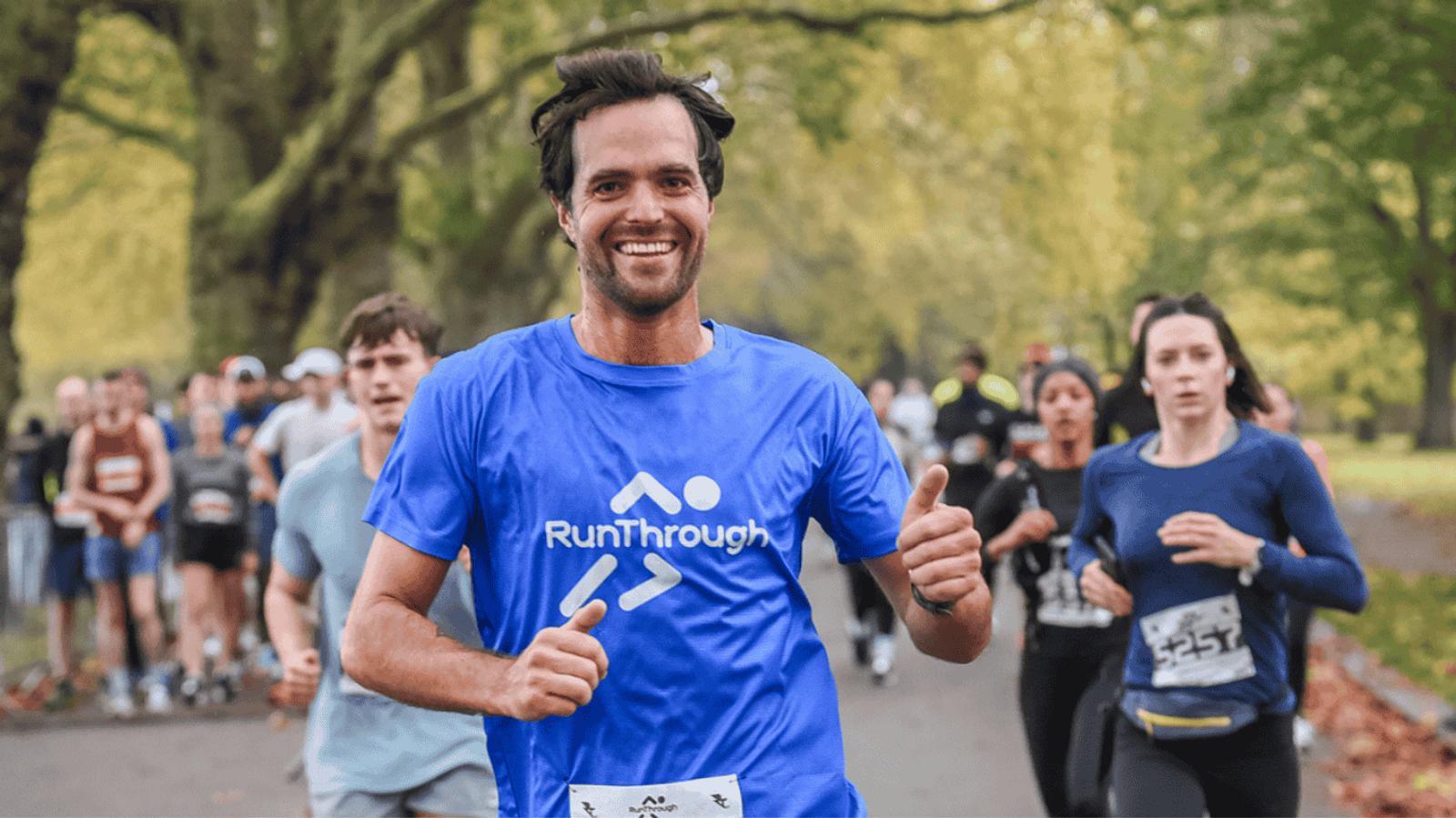 A man in a blue "RunThrough" shirt runs in a park, smiling and giving a thumbs-up. Other runners are visible in the background amid trees with green leaves. It looks like an organized race event.