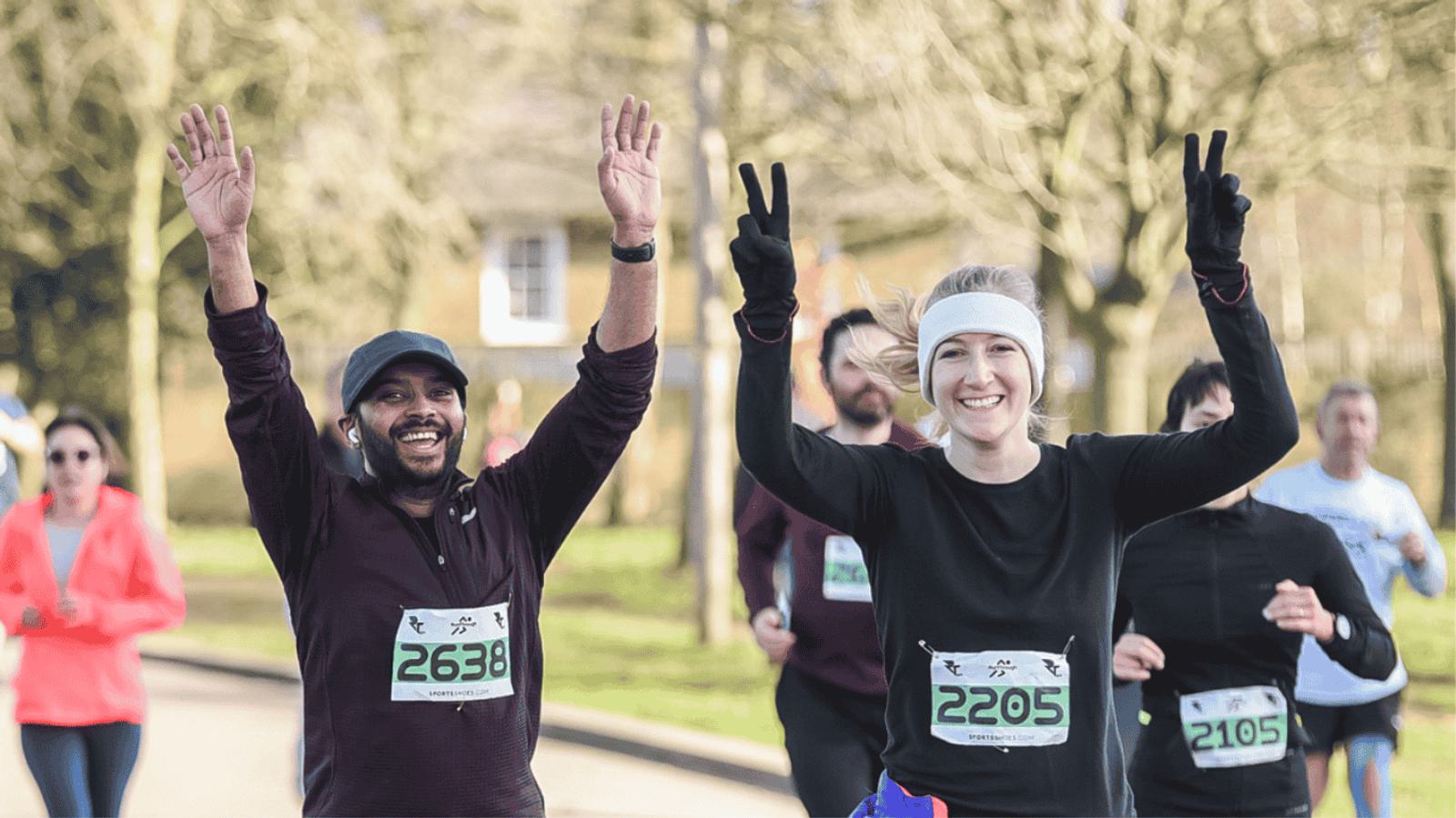 Participants in a running event, wearing race bibs, smile and raise their arms in celebration. They are running on a pathway lined with trees on a sunny day, with other runners in the background.