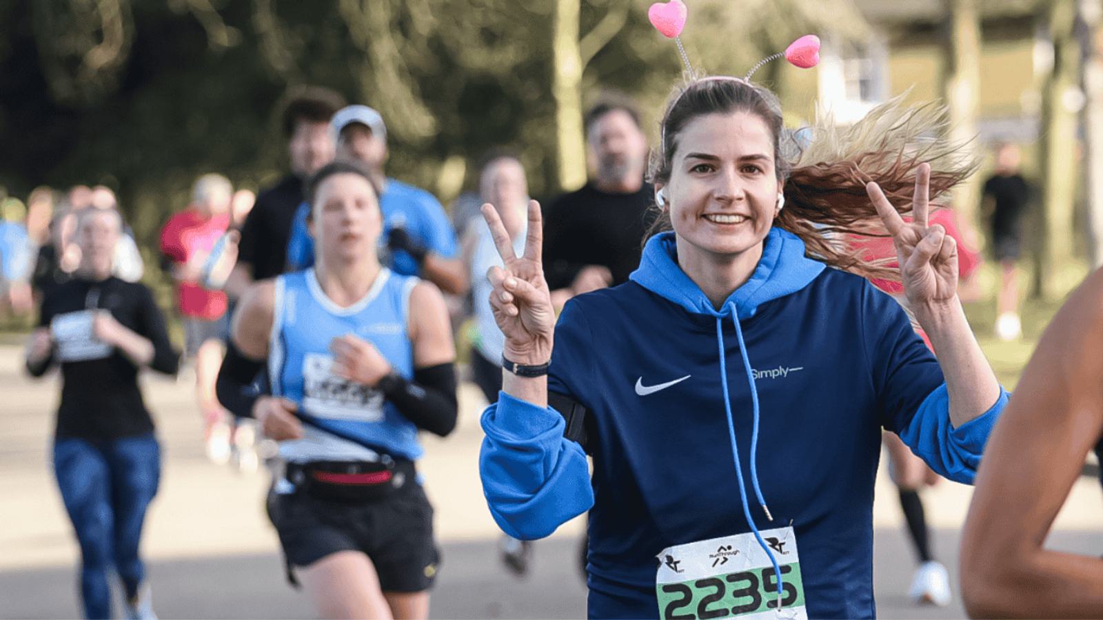 A woman in a blue hoodie with race number 2235 smiles and makes peace signs with both hands while running. She wears pink heart-shaped headbands. Other runners are visible in the background on a sunny day.