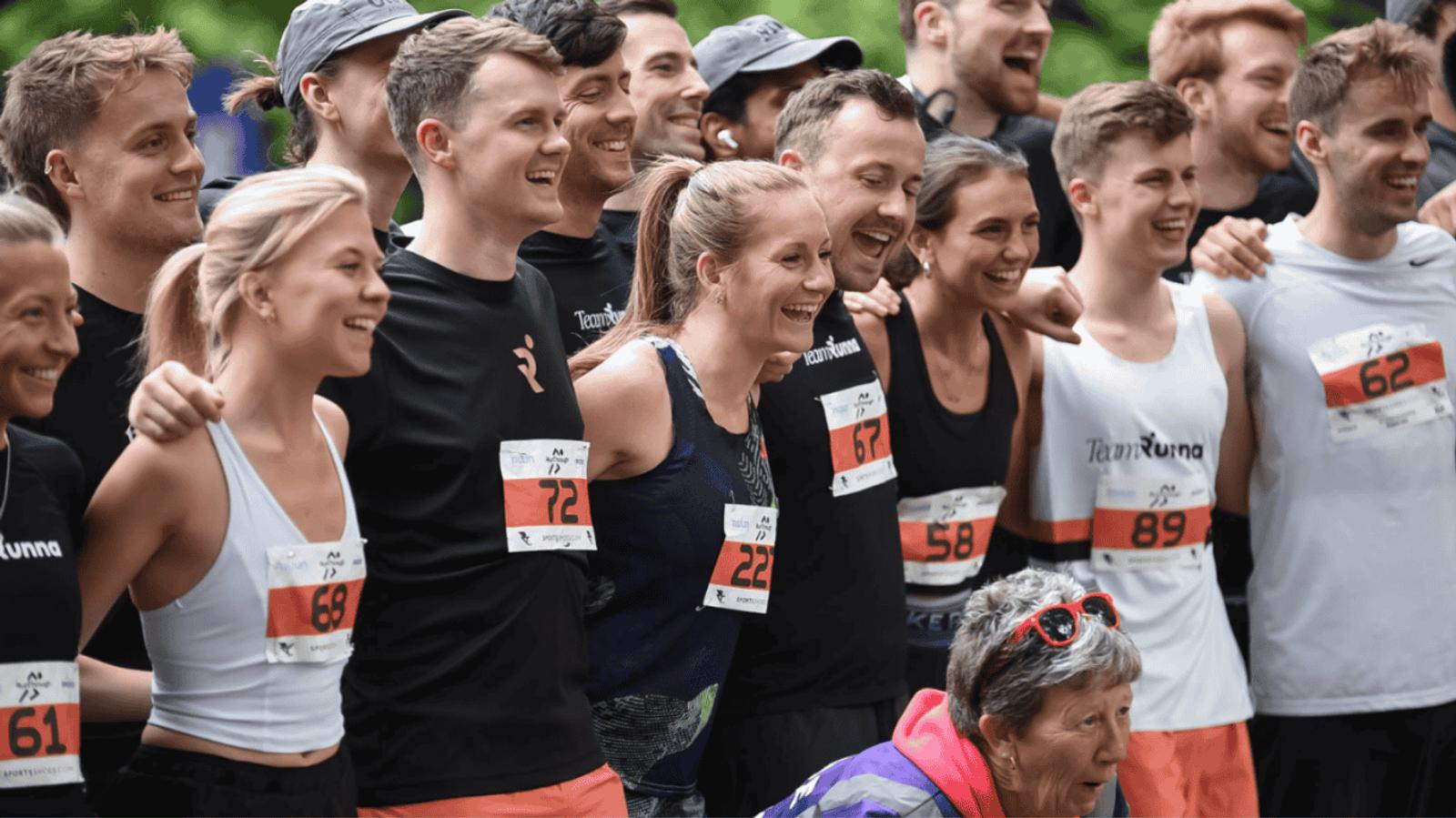 A group of runners, wearing numbered bibs, pose together at an outdoor event. They're smiling and embracing, suggesting a friendly and celebratory atmosphere. A woman in a purple jacket is kneeling in front, also smiling.