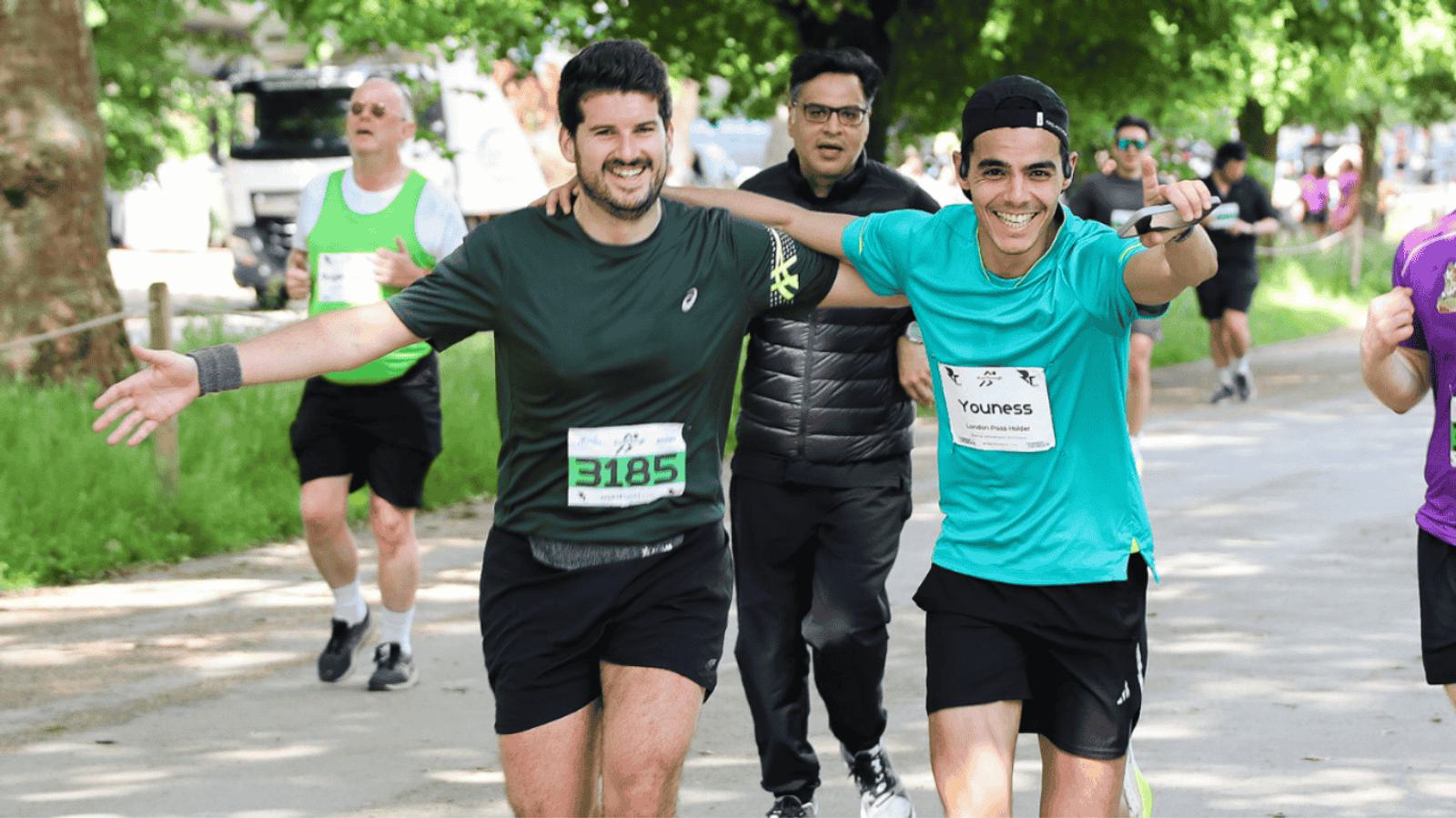 Two men are running in a park, arms around each other, smiling at the camera. Both are wearing numbered bibs and sportswear. Other runners are in the background on a tree-lined path.