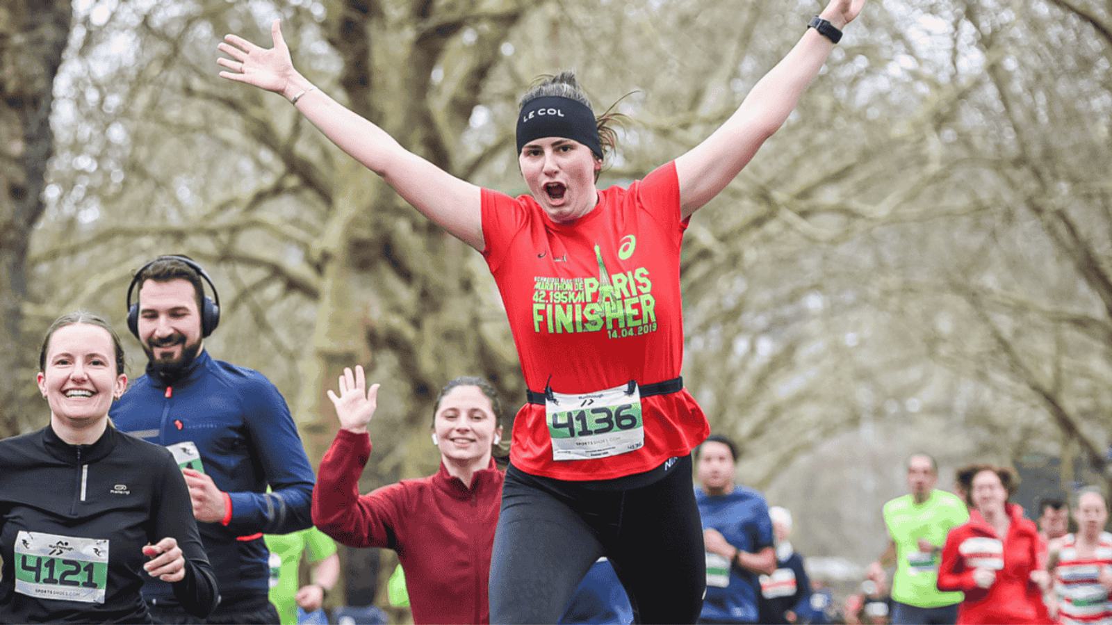 A runner wearing a red shirt and black pants celebrates with arms raised crossing the finish line in a race. Other runners are visible in the background, some smiling, with trees lining the path.