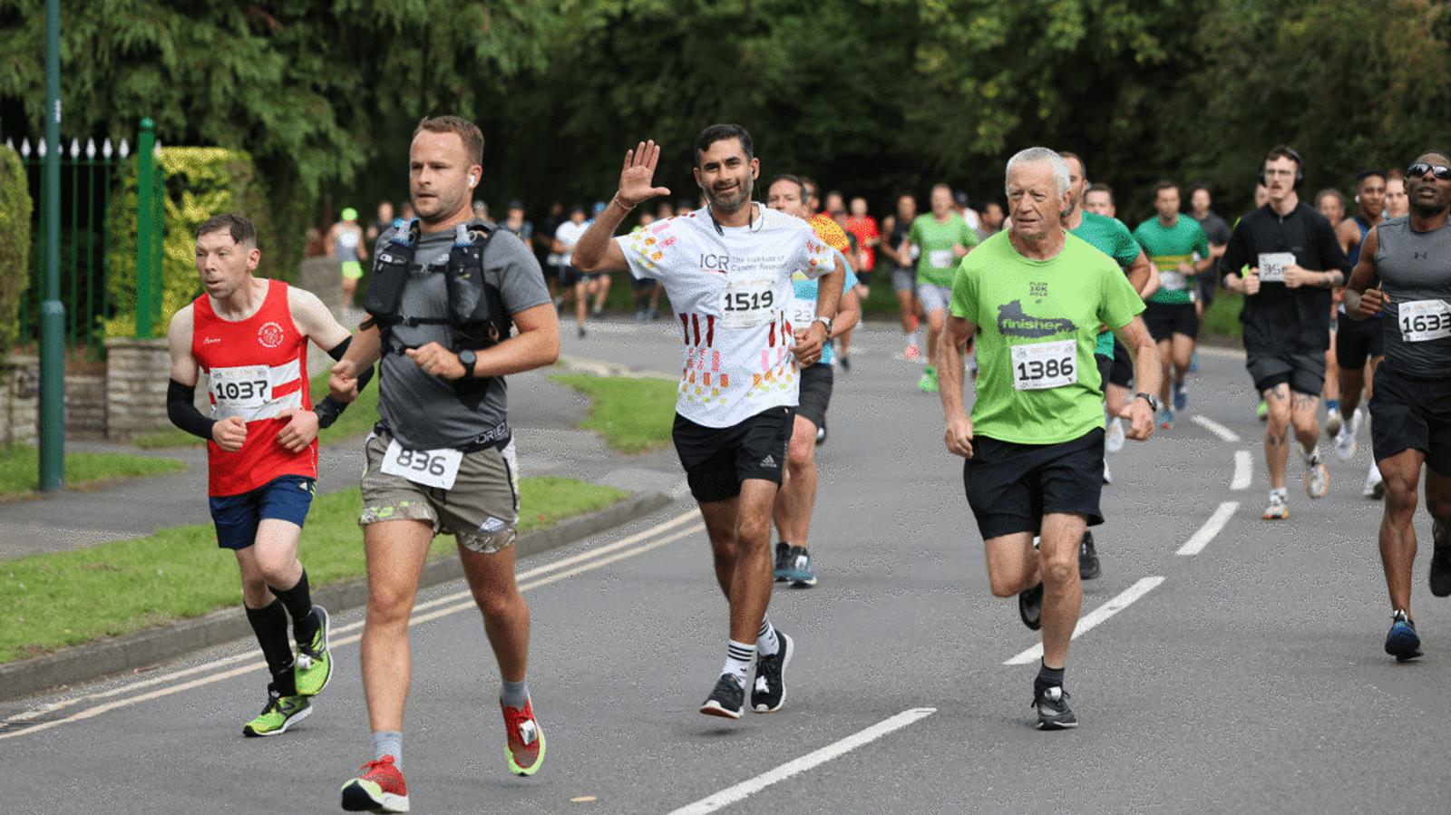 A group of runners participating in a race on a road. The lead runner wears a white bib numbered 836; another smiles and waves, wearing bib 1519. The greenery in the background suggests a park or wooded area.