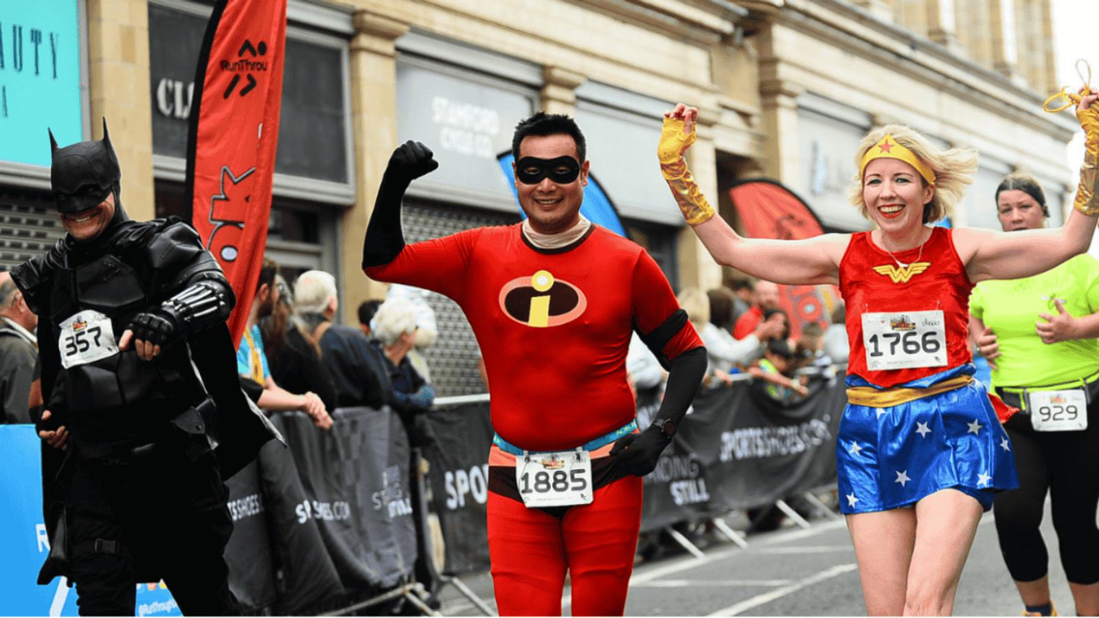 Three runners participate in a race wearing superhero costumes: one as Batman, another as Mr. Incredible, and the third as Wonder Woman. They are smiling and raising their arms in celebration as they run past cheering spectators.