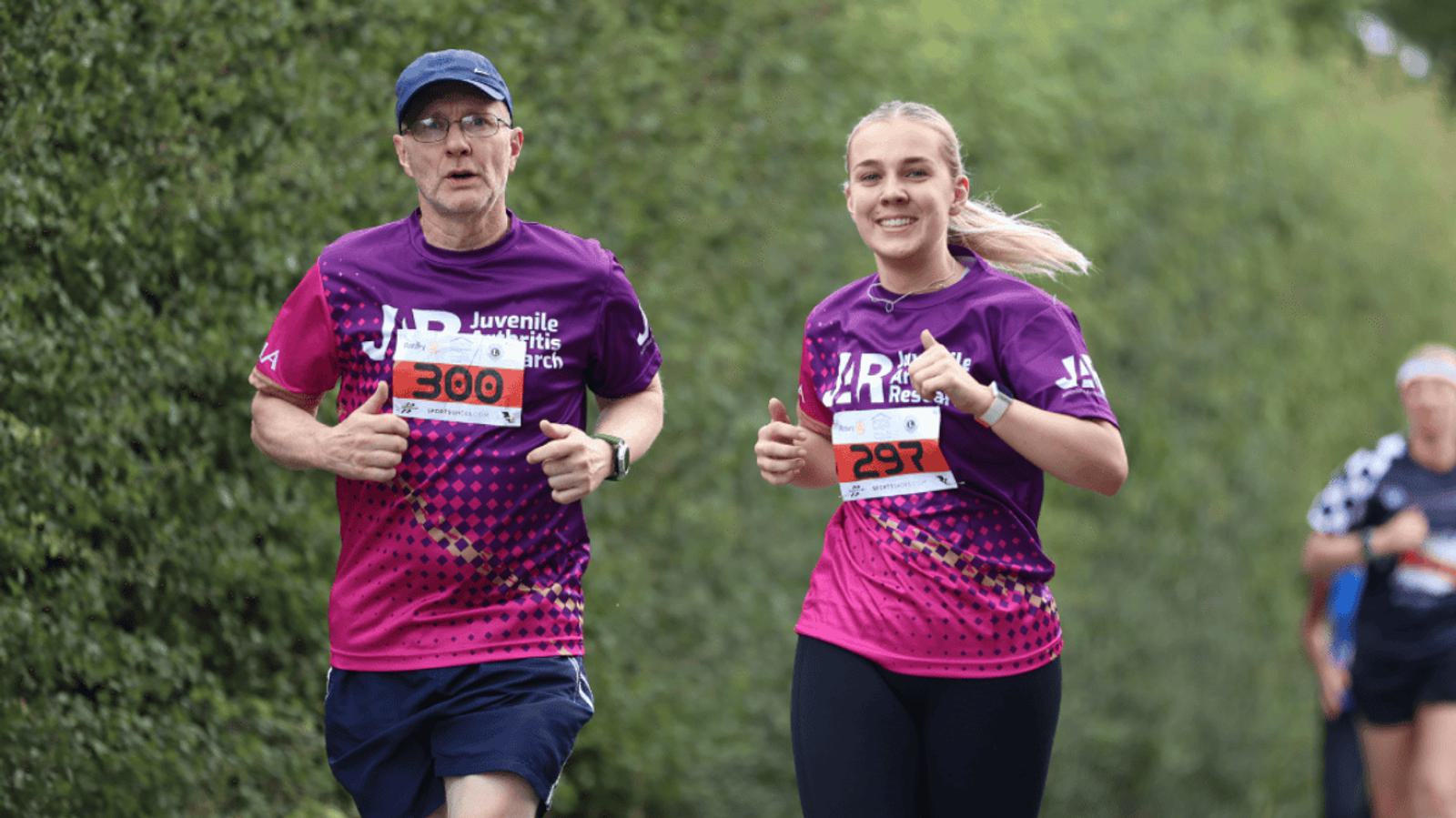 Two runners in purple shirts, marked with numbers 300 and 297, smile and give thumbs up while jogging along a tree-lined path. Other runners are visible in the background.