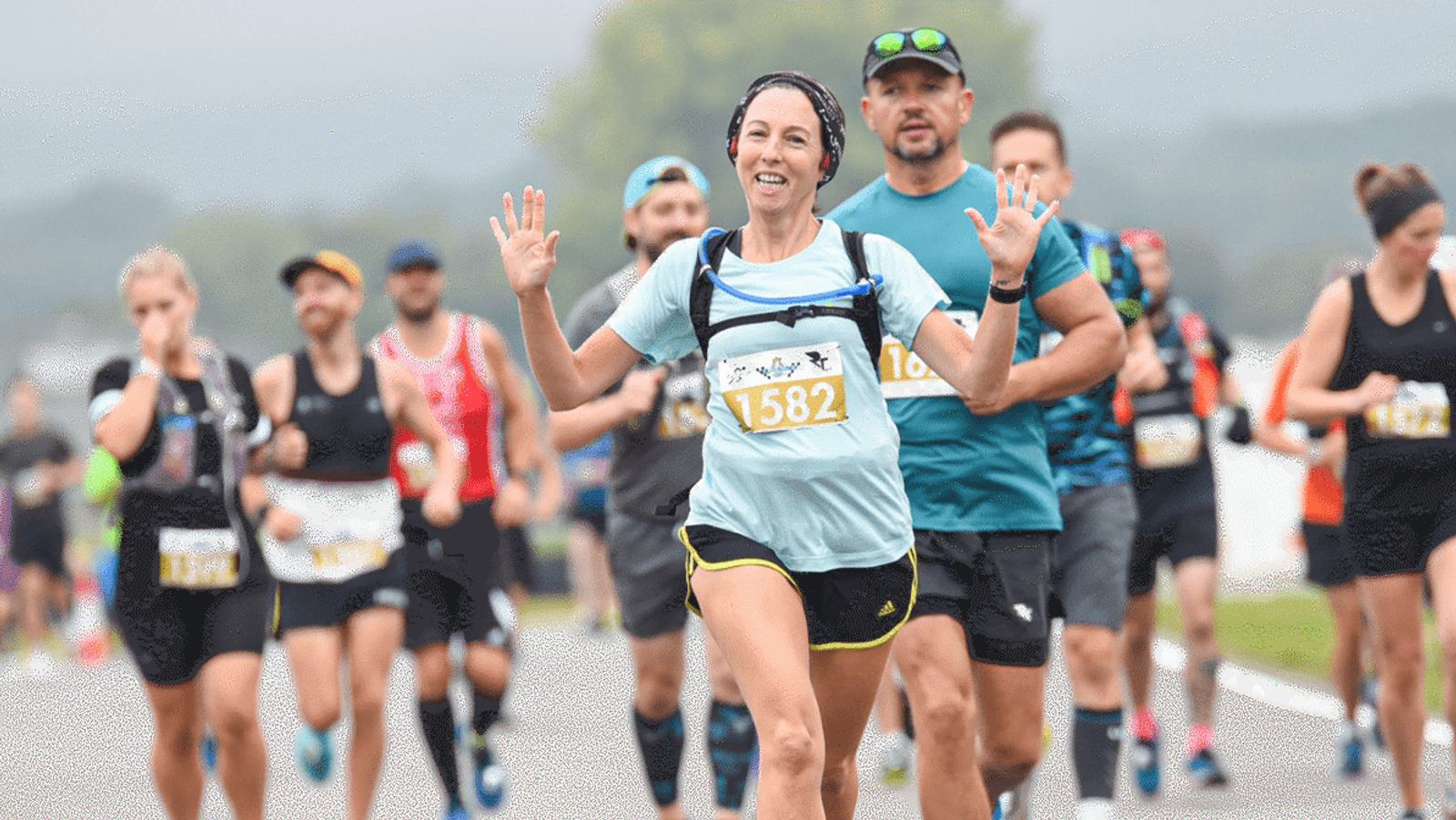 A group of runners participating in a race. A woman in the foreground, wearing a light blue shirt and a yellow race bib numbered 1582, is smiling and raising her hands. Other runners are visible in the background on a road.