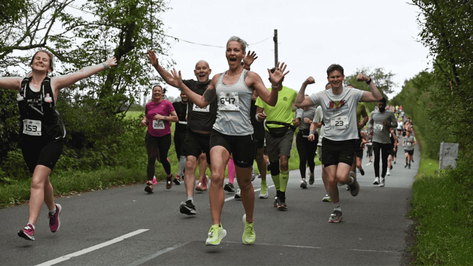 A group of runners is happily participating in a road race. They are surrounded by green trees and appear joyful, raising their arms in excitement. The sky is overcast, and the road is lined with lush greenery.