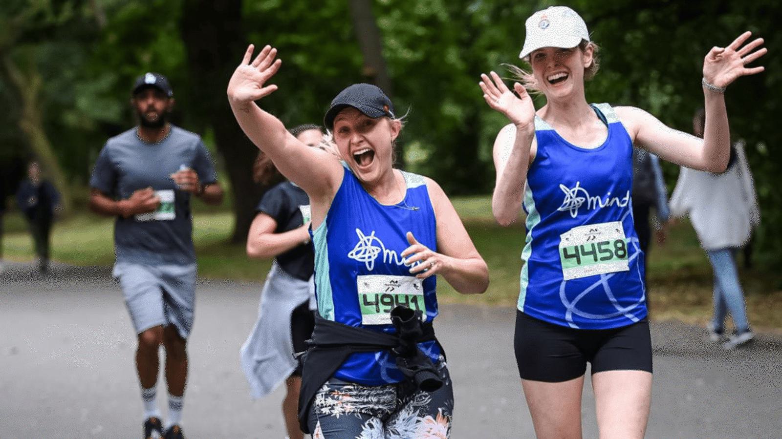 Two women in blue athletic gear smile and wave at the camera while running in a park. They each wear numbered race bibs. A man and another person follow behind. The setting is lush and green, suggesting a pleasant day for a run.