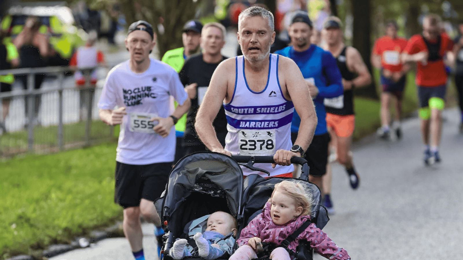 A man running a marathon pushes a stroller with two young children. He's wearing a "South Shields Harriers" singlet. Other runners are visible in the background, some wearing race numbers. The setting is along a road with greenery.
