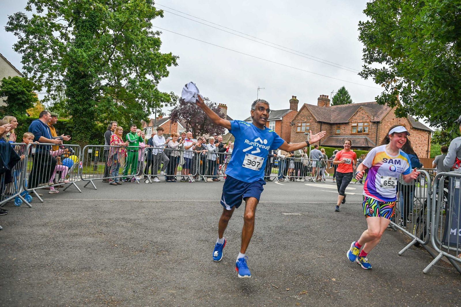Two runners, smiling broadly, approach the finish line during a race. One holds a shirt in the air. Spectators cheer behind a barricade under overcast skies. Nearby buildings and trees border the street.