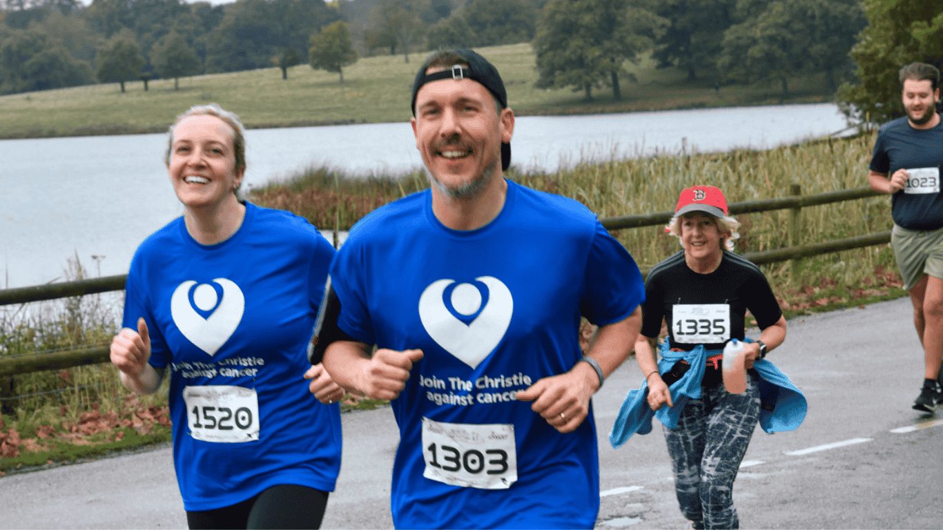 Three people in blue T-shirts with white heart logos run in a park during an event, smiling. Another person follows, holding a drink. The background shows trees and a lake. The T-shirts read, "Join The Christie Against Cancer.