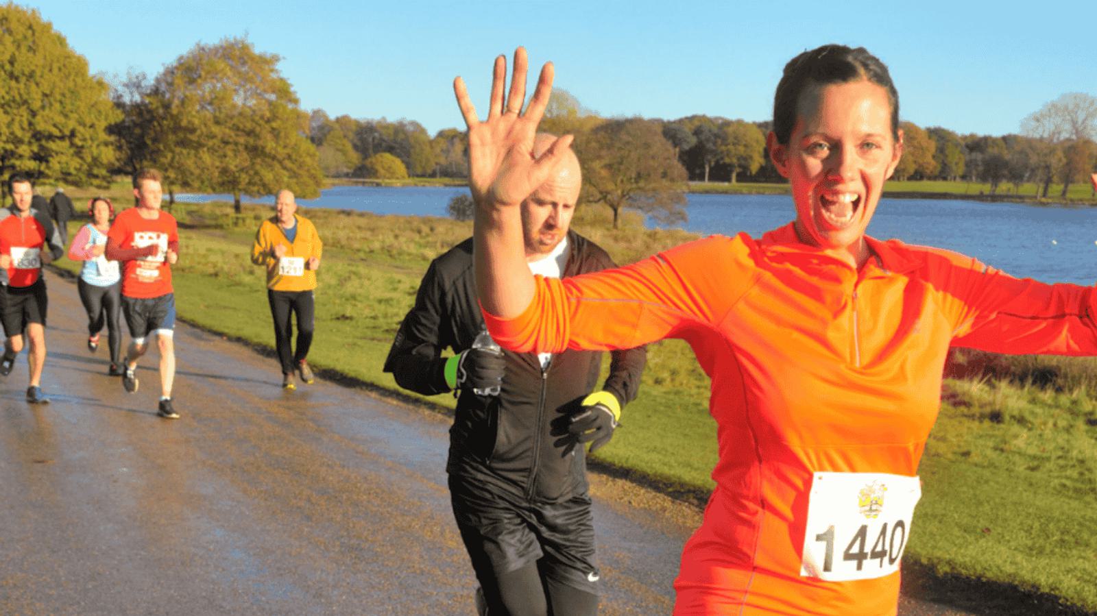 A woman in a bright orange running outfit smiles and waves at the camera while running along a lakeside path. Several other runners are visible in the background on a sunny day with trees and a lake in view.