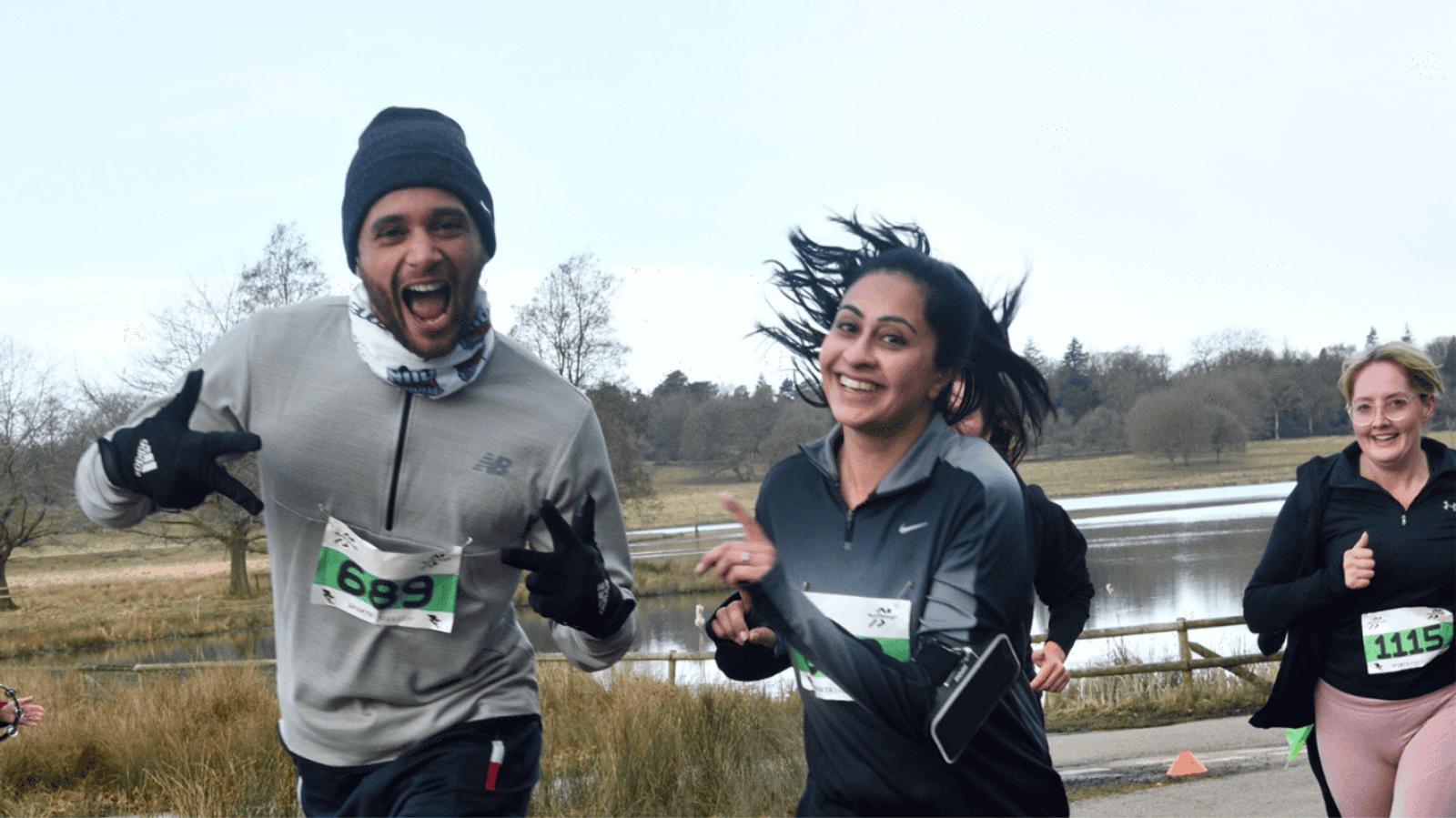 Three people are running in a race on a path beside a lake. The man on the left is wearing a beanie and neck gaiter, smiling with his thumbs up. The two women, one of whom wears a headband, are also smiling. They are all wearing numbered bibs.