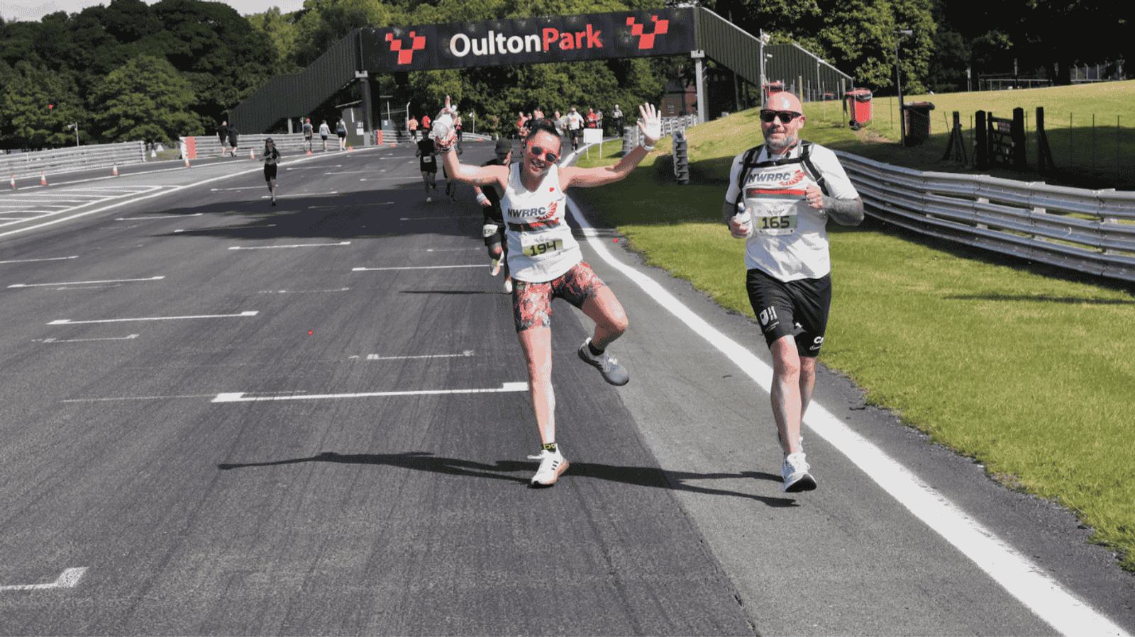 Two runners celebrate as they approach the finish line at Oulton Park. They are wearing athletic gear and sunglasses. The road is lined with spectators and a bridge with the Oulton Park sign spans the track above them.
