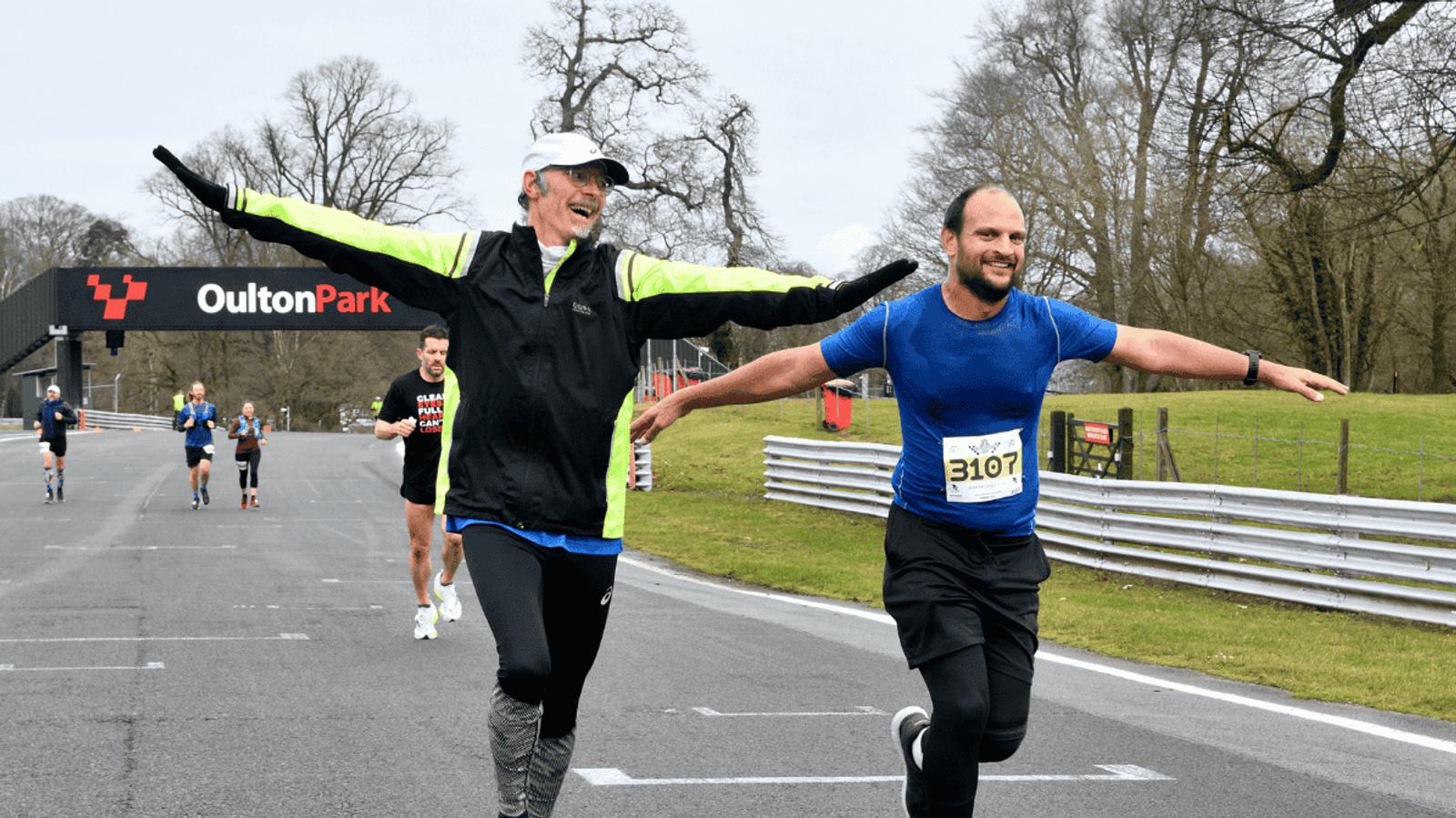 Two men are running on a racetrack with arms outstretched, smiling broadly. One wears a yellow and black jacket, the other a blue shirt. In the background, other runners and the Oulton Park sign are visible. Trees and grass line the track.