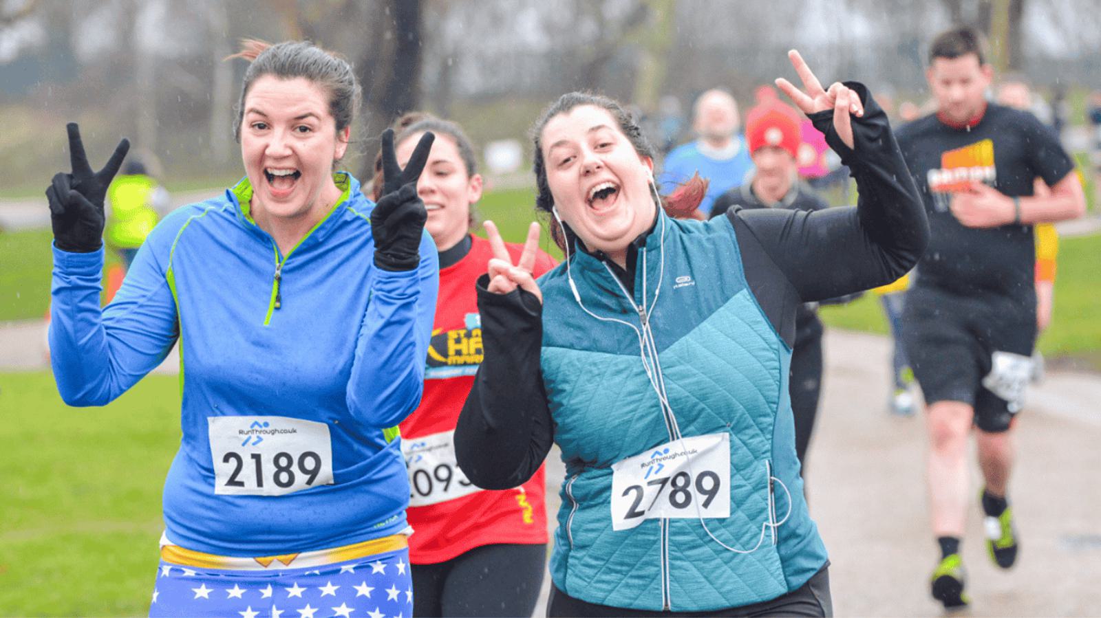 Two women wearing race numbers 2189 and 2789 smile and make peace signs as they run in an outdoor event. Other participants are jogging behind them on a path surrounded by grass. The weather appears overcast.