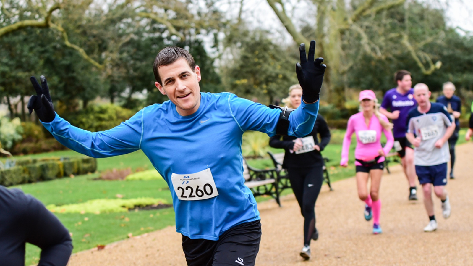 A group of runners in a park, led by a man in a blue shirt with race number 2260. He is smiling and gesturing a peace sign. Other runners follow behind, wearing different athletic gear. Lush green trees and a pathway are visible in the background.