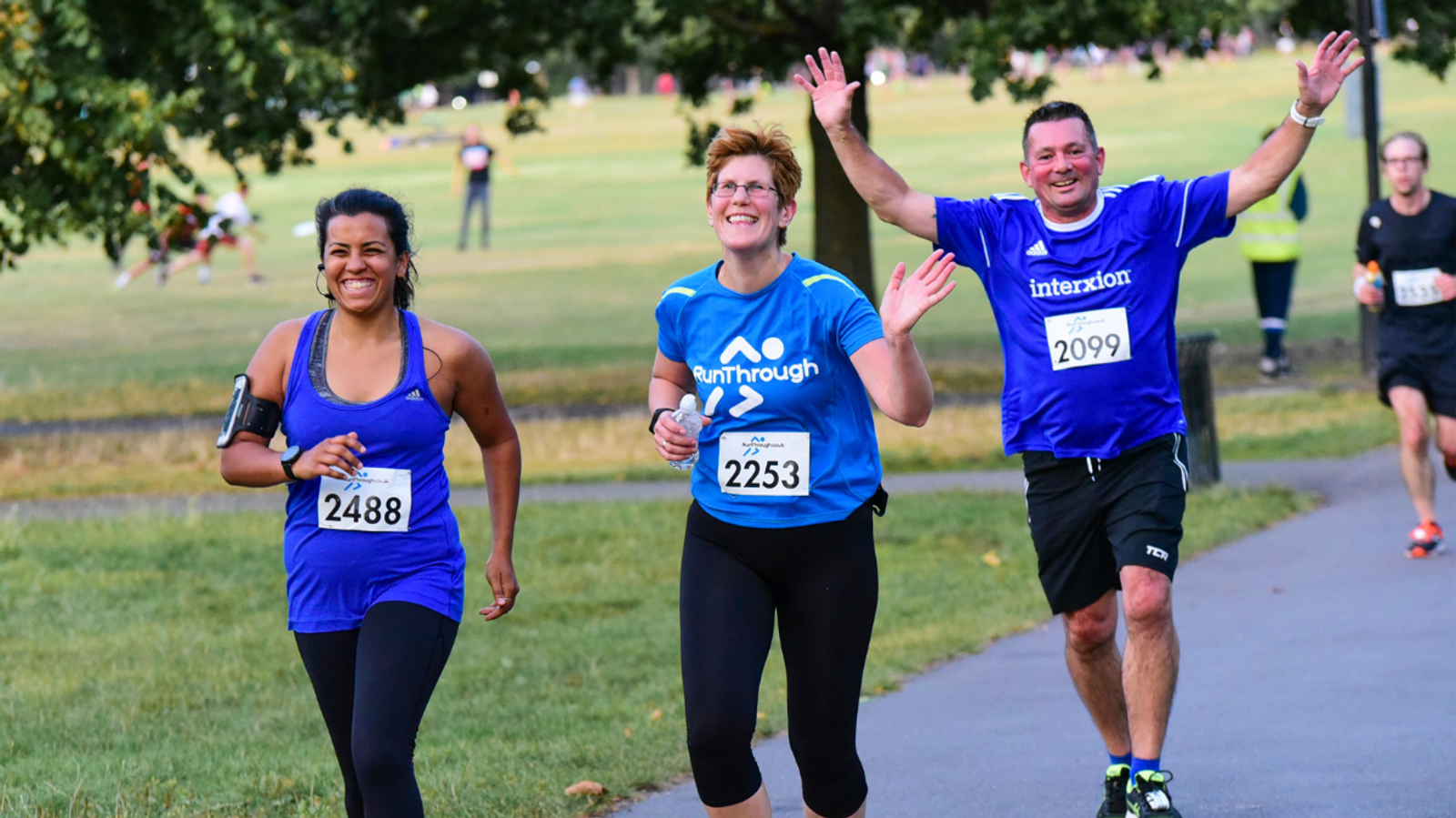 Three runners in blue shirts jogging and smiling in a park. Two women and one man, with race numbers visible: 2488, 2253, and 2099. The path is surrounded by grass and trees.