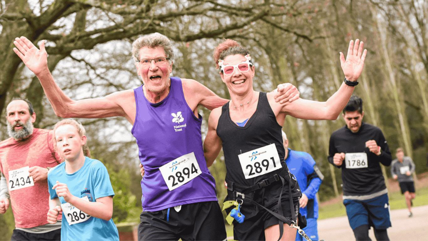 A group of runners, including a smiling elderly man in a purple tank top and a joyful woman in sunglasses, proudly pose mid-run with race numbers 2894 and 2891. They are in a park setting with trees in the background.