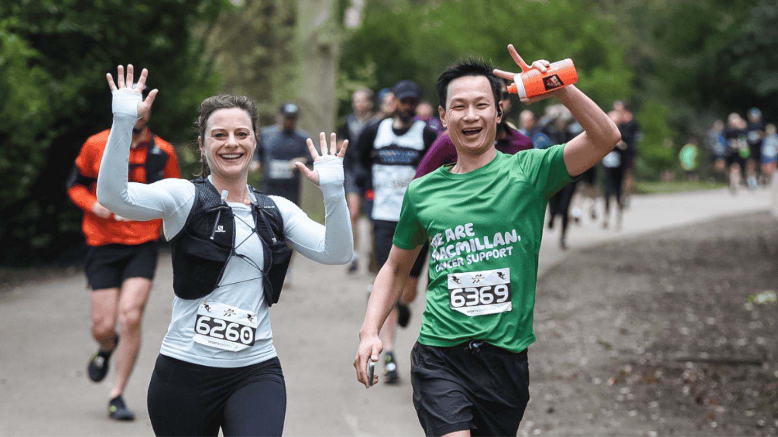 Two runners smile and wave at the camera during a race. The woman wears a white long-sleeve top and the man wears a green shirt with "Macmillan Cancer Support." Other runners are visible in the background on a tree-lined path.