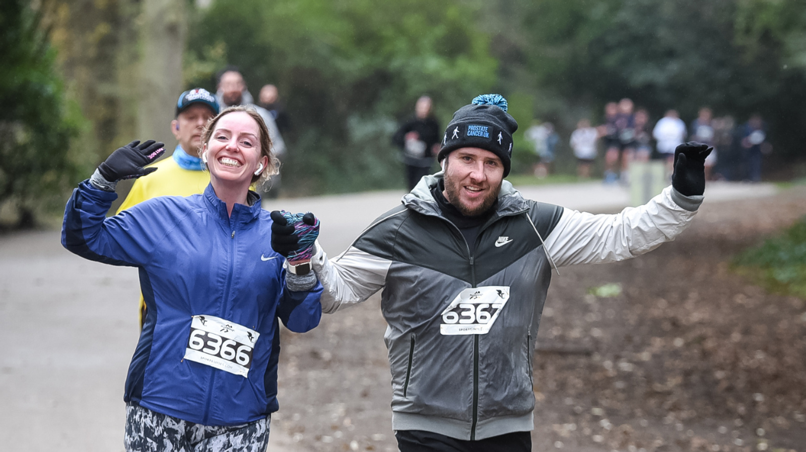 Two people wearing numbered bibs, a woman and a man, run on a park path, smiling and raising their arms. They are dressed in warm athletic wear. Other runners can be seen in the background.