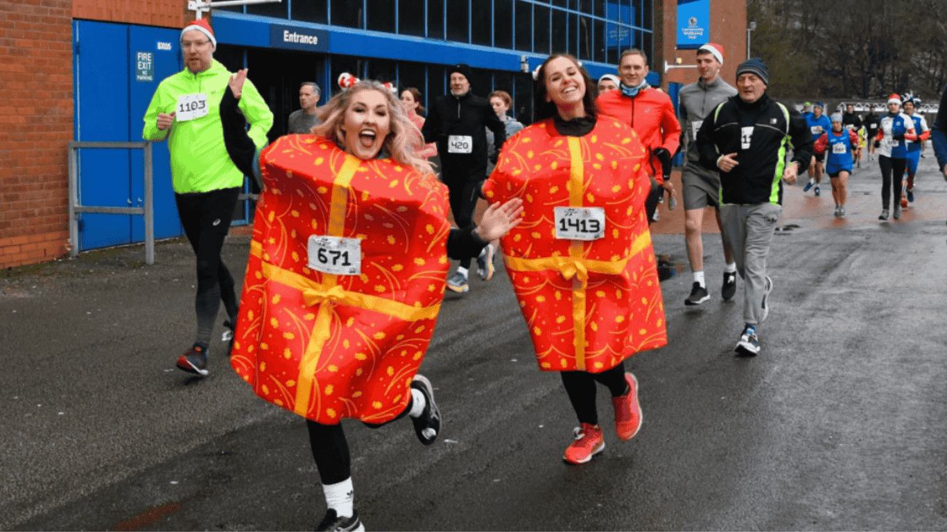 Two runners in red and yellow gift-wrapped costumes, with race numbers 671 and 143, smile and jog during a fun run event. Other participants in athletic gear are behind them, near a building labeled "Entrance." The ground is wet, indicating recent rain.