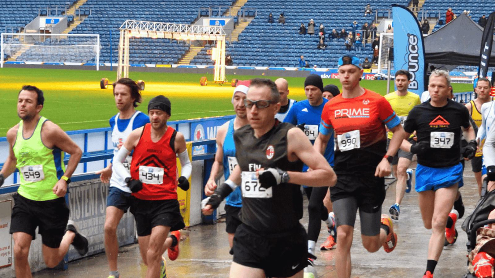 A group of runners participating in a race on a track in a stadium. The participants are wearing numbered bibs and a variety of athletic gear. The seating area is visible in the background, with a few spectators watching.