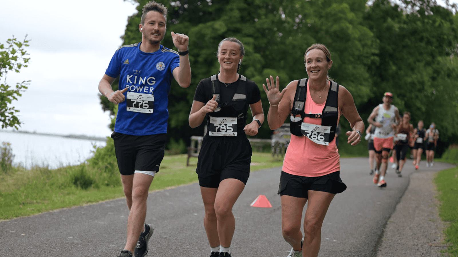 Three people are jogging and smiling on a paved path surrounded by greenery. Two women are wearing race bibs 785 and 786, and a man is wearing a bib with the number 776. Other runners can be seen in the background.