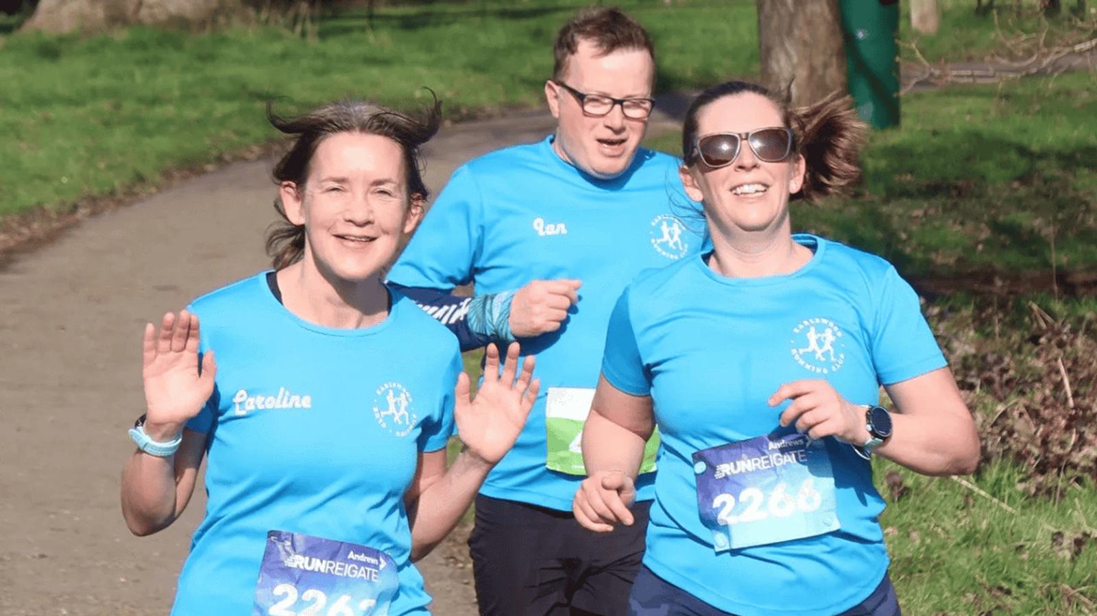 Three people in blue running shirts participate in a race. The woman on the left waves, and they are jogging on a path with greenery around them. Each has a race bib with numbers, and they appear to be enjoying the run.