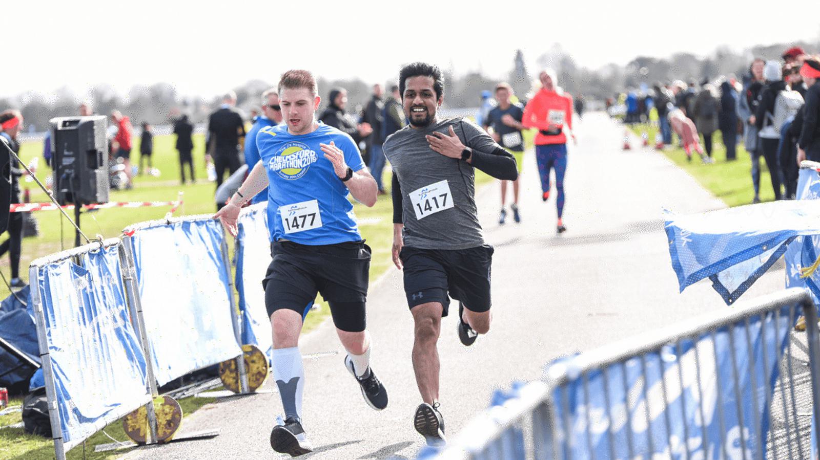 Two runners sprint towards a finish line on a sunny day, with one wearing a blue shirt and the other in a gray and black outfit. Both have race bibs, with onlookers and other runners in the background.