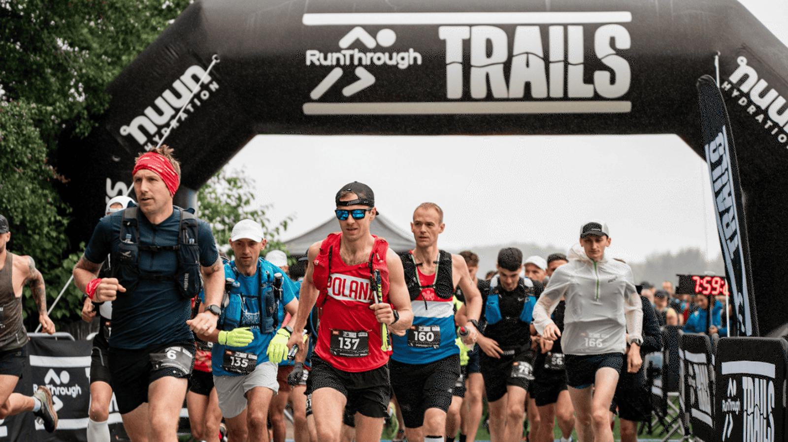 A group of runners begins a race under a black arch with "RunThrough Trails" written on it. The lead runner wears a red jersey with "Poland" on it. Other runners in various athletic gear and hats follow closely. Trees and a digital clock are visible.