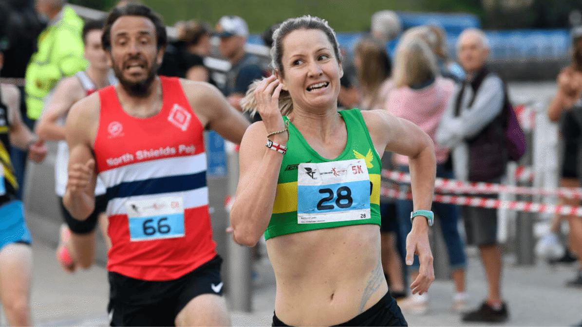 A woman in a green and yellow tank top, bib number 28, smiles and runs a race, with a male runner in a red, white, and blue tank top, bib number 66, close behind. Spectators and a barricade are visible in the background.