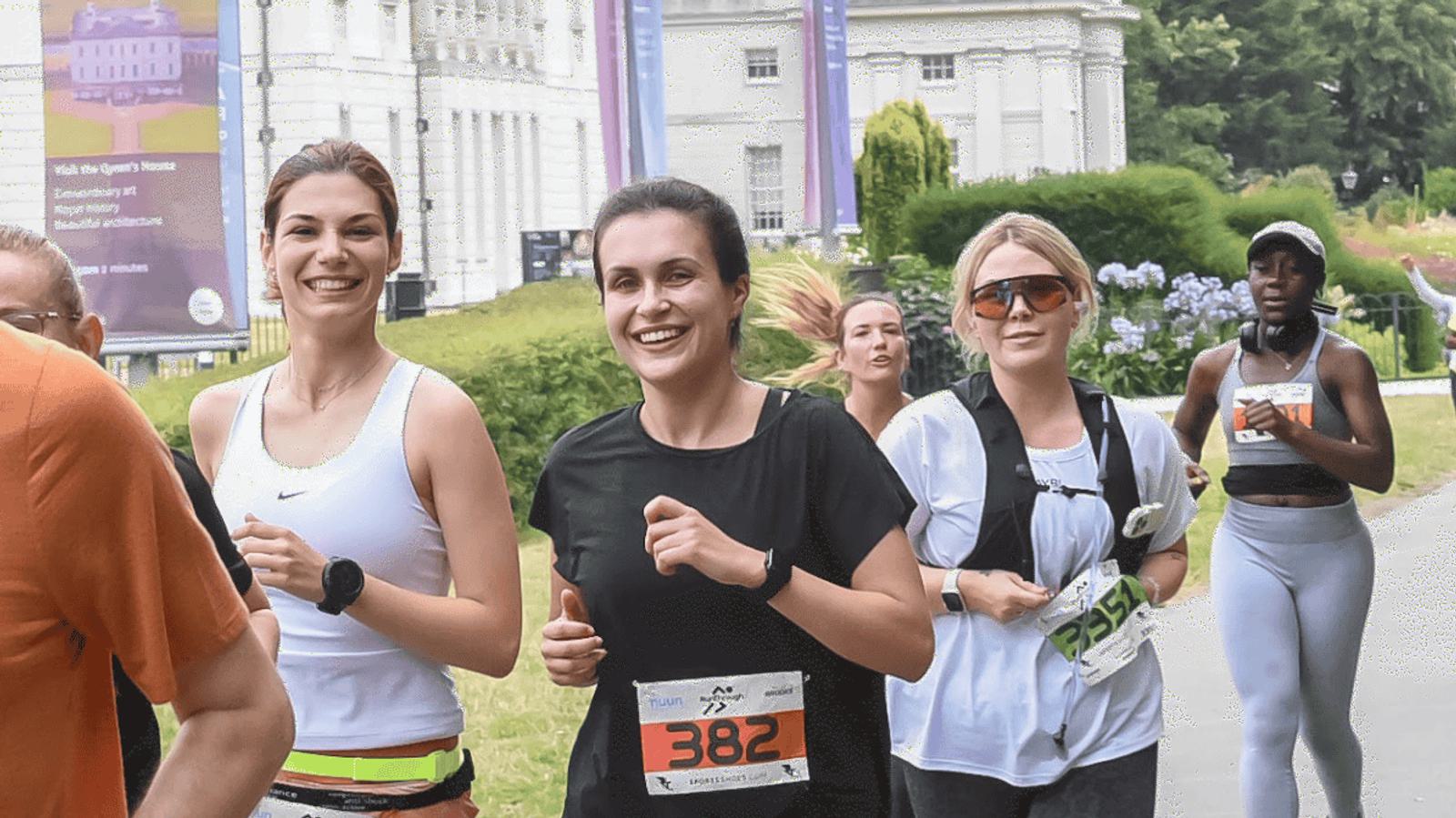 A group of runners participating in a race, smiling as they pass a park. Two women in the foreground wear race bibs numbered 382 and 384. Participants are dressed in athletic gear, with greenery and a building in the background.