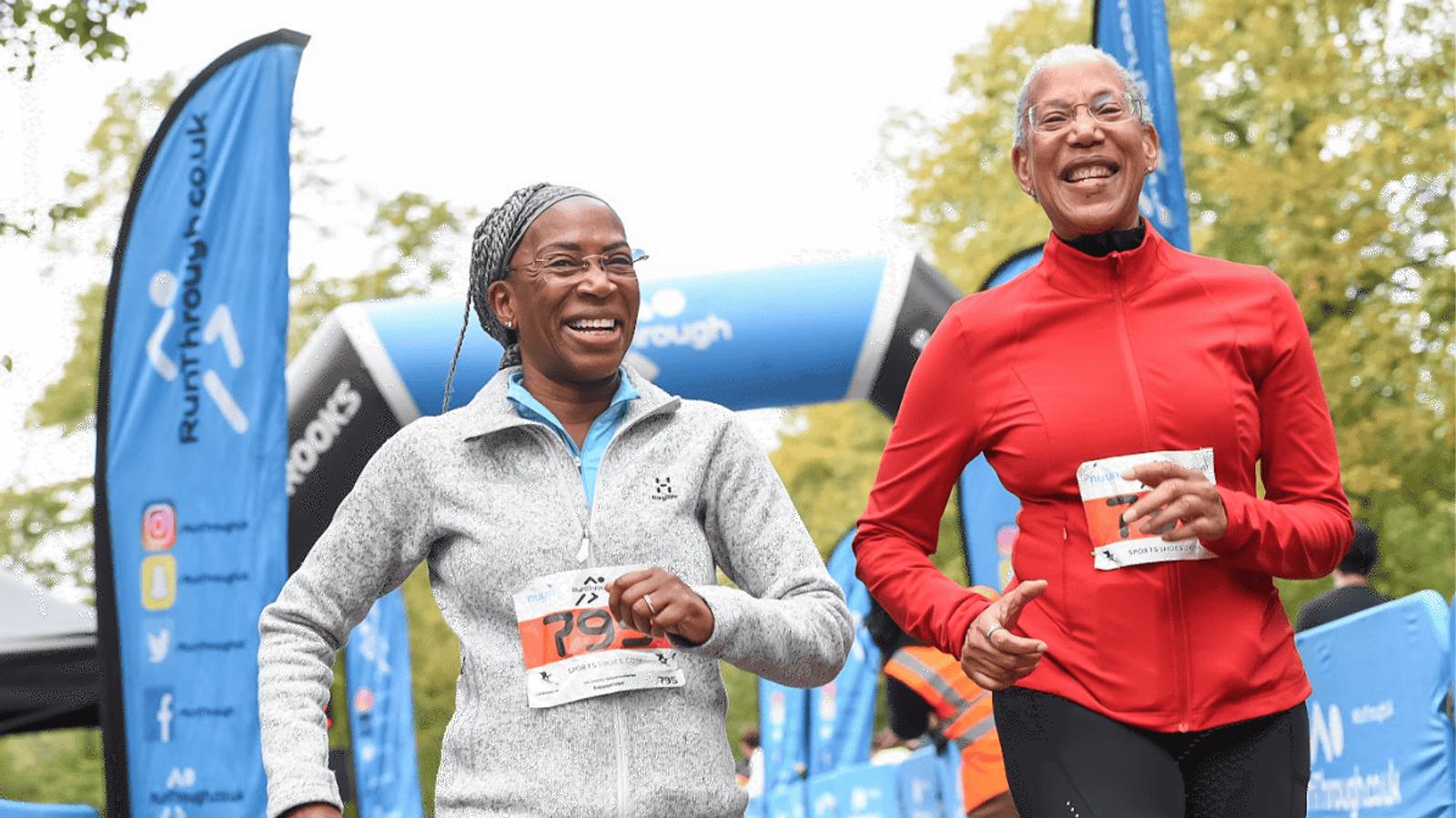 Two people in athletic gear smile as they run in a race event. One wears a gray jacket and glasses, the other a red jacket. Blue banners and an inflatable arch are visible in the background. They both carry race numbers.