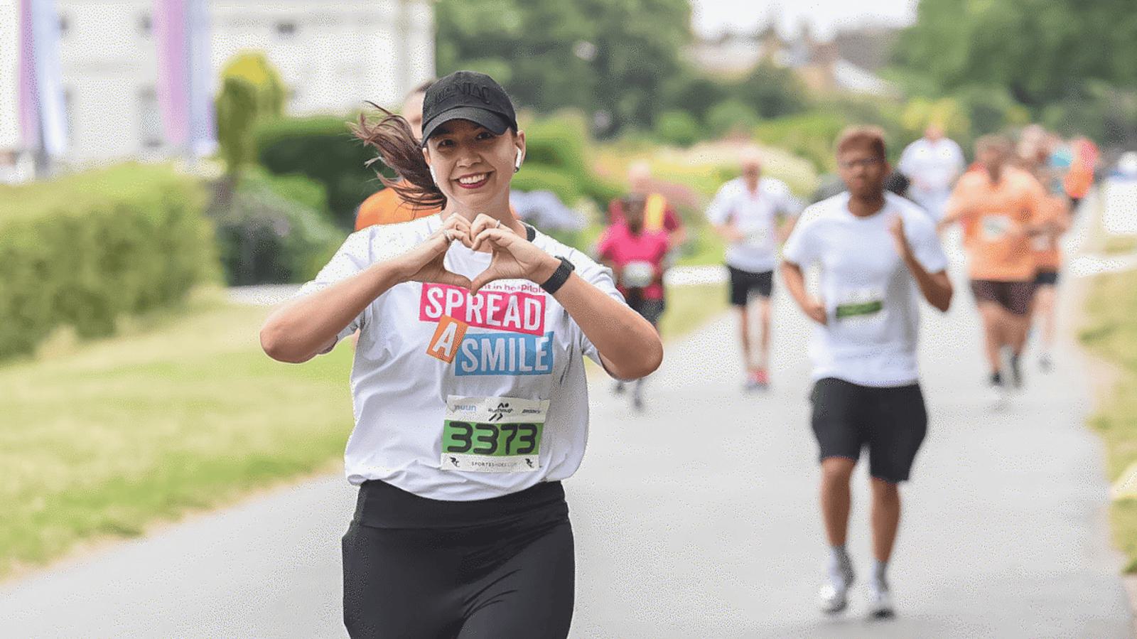 A woman in athletic gear runs a race, smiling and making a heart shape with her hands. Her shirt reads "Spread 4 Smile." Other runners are visible in the background on the tree-lined path.