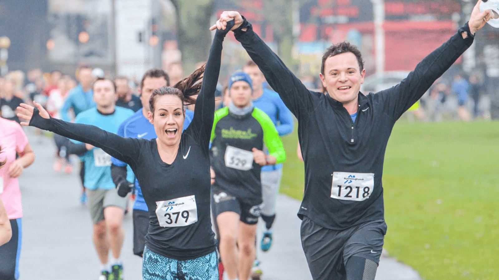 Two runners, a man and a woman, joyfully raise their arms while holding hands during a race. Both wear race numbers, and other participants can be seen behind them on the path. The event takes place on a cloudy day.