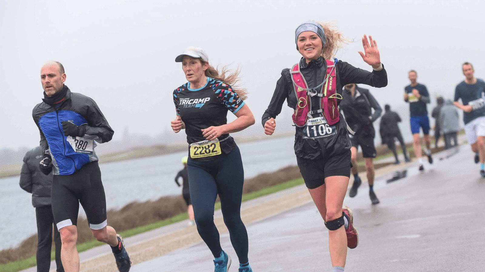 Runners on a wet path during a race. A woman in black with a pink vest waves, another wears a white cap, and a man in black runs nearby. Others follow behind, with a lake and overcast sky in the background. Bib numbers are visible.