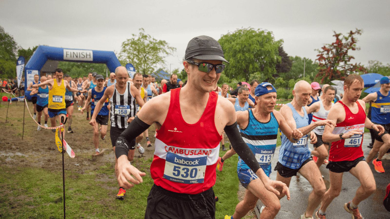 Runners in athletic wear begin a race under a blue "Finish" inflatable archway, despite it being the start. The ground is wet, indicating recent rain. The front runner wears a red vest and black cap, smiling as others follow closely behind. Trees line the path.