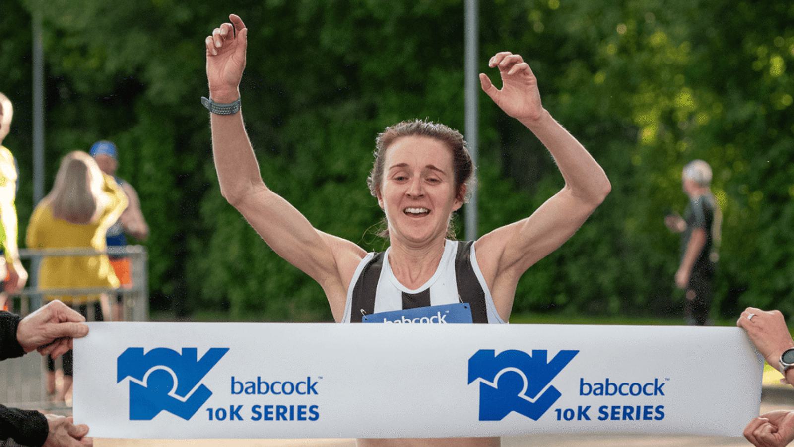 A runner crossing the finish line with arms raised in victory at the Babcock 10K Series. The athlete is wearing a black and white tank top. Blurred spectators and greenery are visible in the background.